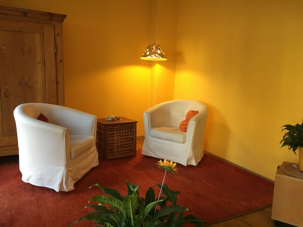 Zwei weiße Stühle auf einem roten Teppich in ambientischem Licht. Es stehen Pflanzen, ein Schrank und ein Tisch im Raum.